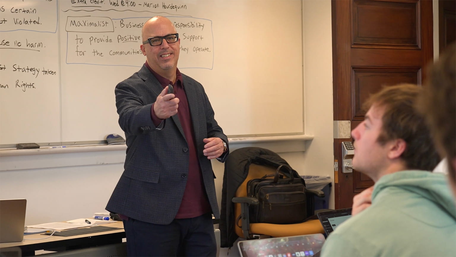 Professor Caruso in a blazer pointing toward a student during a classroom lecture with notes on a whiteboard behind him.