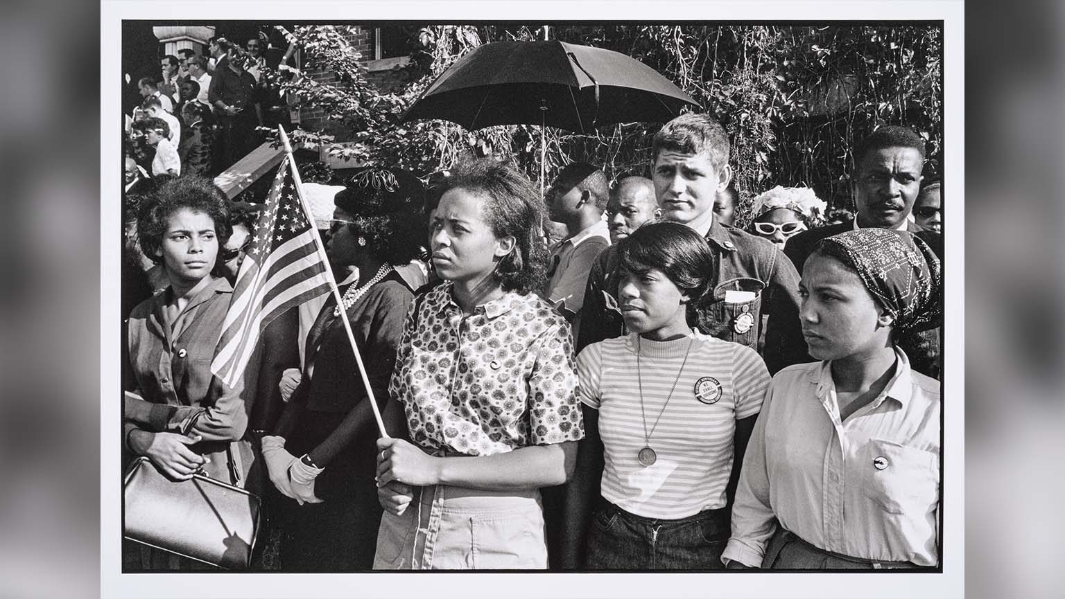 A black and white photograph by Danny Lyon titled “SNCC workers stand outside the funeral: Emma Bell, Dorie Ladner, Dona Richards, Sam Shirah, and Doris Derby, Birmingham” depicting a group of solemn young African American individuals, some holding American flags, standing closely together waiting outside a funeral.