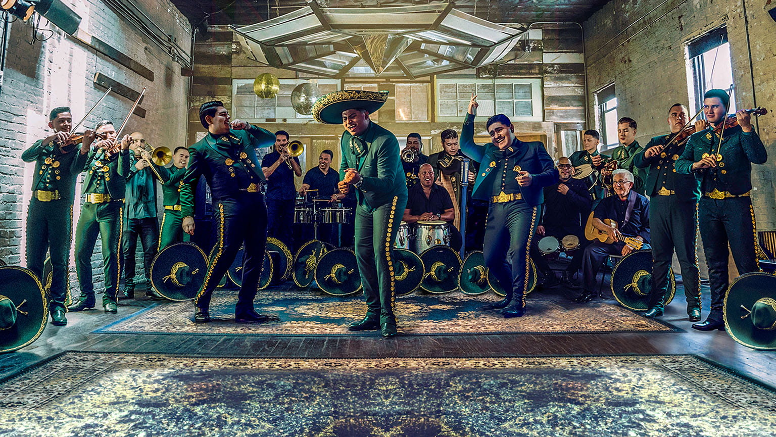 Fourteen-member Latin mariachi band in matching attire performing one of their hits, complete with violins, trumpets, trombones, drums, and an iconic sombrero hat.