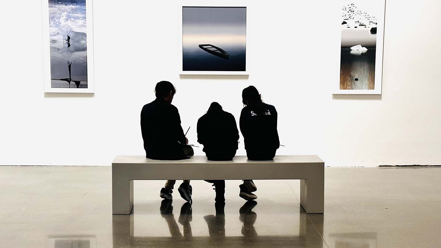 Three people relax on a bench, chatting in front of a wall adorned with various pictures.