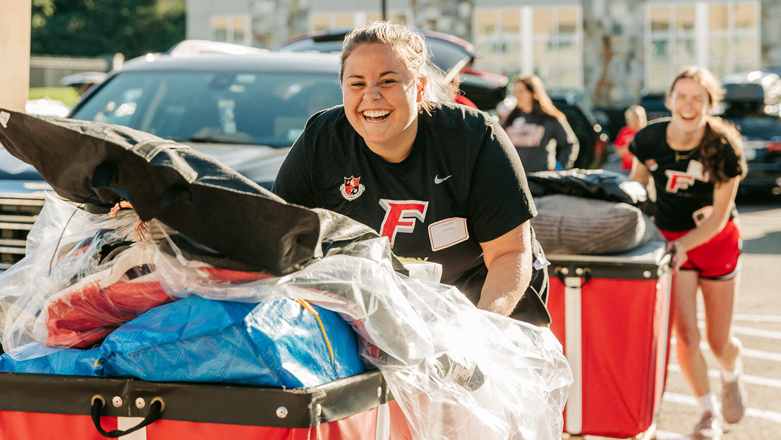 Smiling volunteer in a black F-shirt pushes boxes in a cart as others push red bins outdoors in bright sunlight.