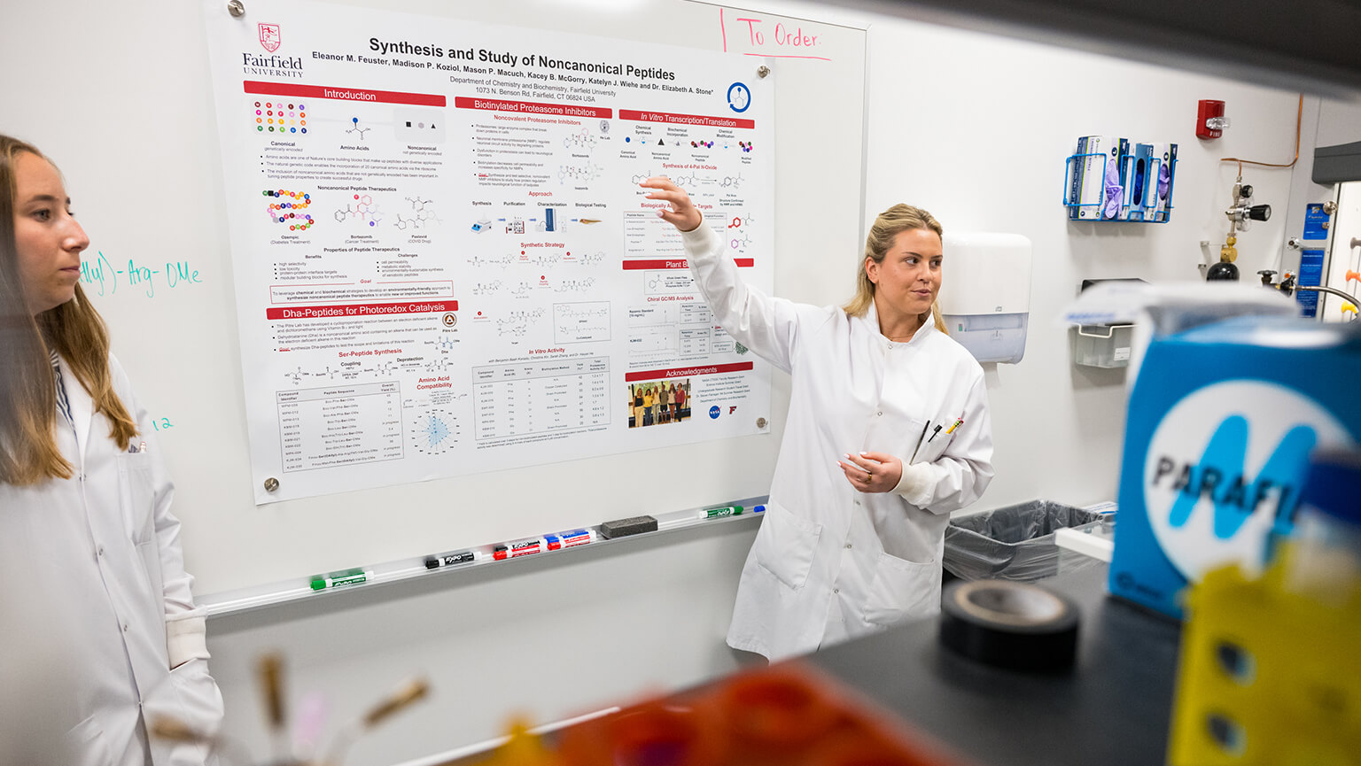 Researchers in white lab coats discuss a scientific poster on noncanonical peptides in a laboratory, creating an informative, collaborative mood.
