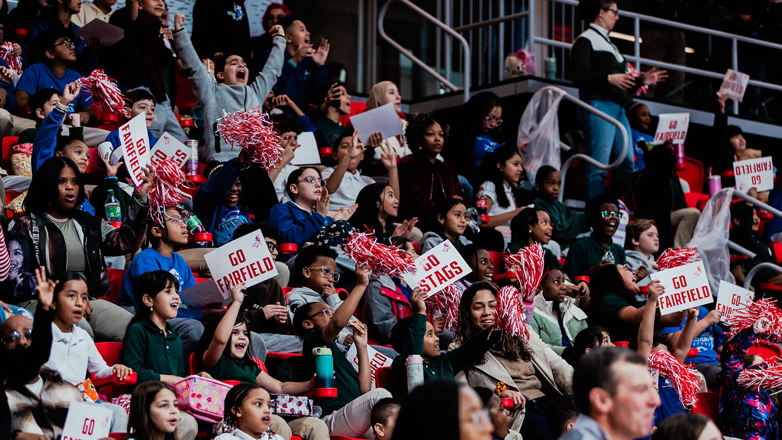 Energetic college sports crowd cheers with red-and-white pom-poms and “Go Fairfield” and “Fairfield Stags” signs in the stands.