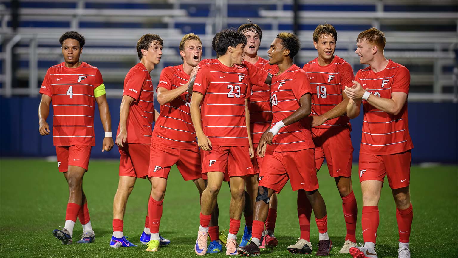 Players from the men's soccer team celebrate a goal, hugging and high-fiving in a moment of joy and camaraderie.