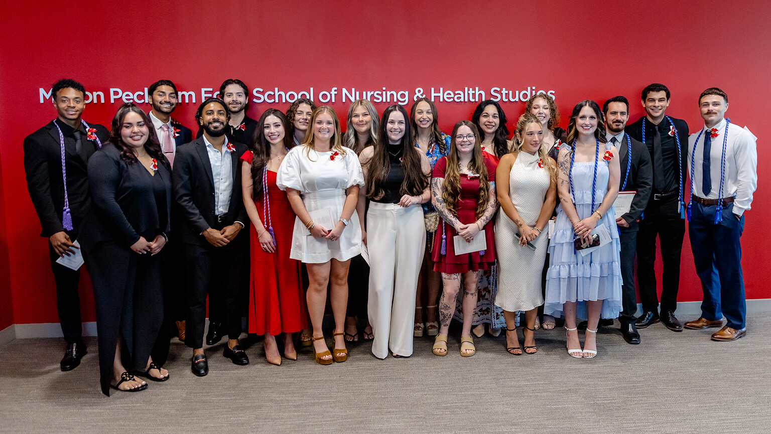 Group photo of diverse nursing and health studies graduates posing in formal attire before a red backdrop, smiling in a celebratory mood.