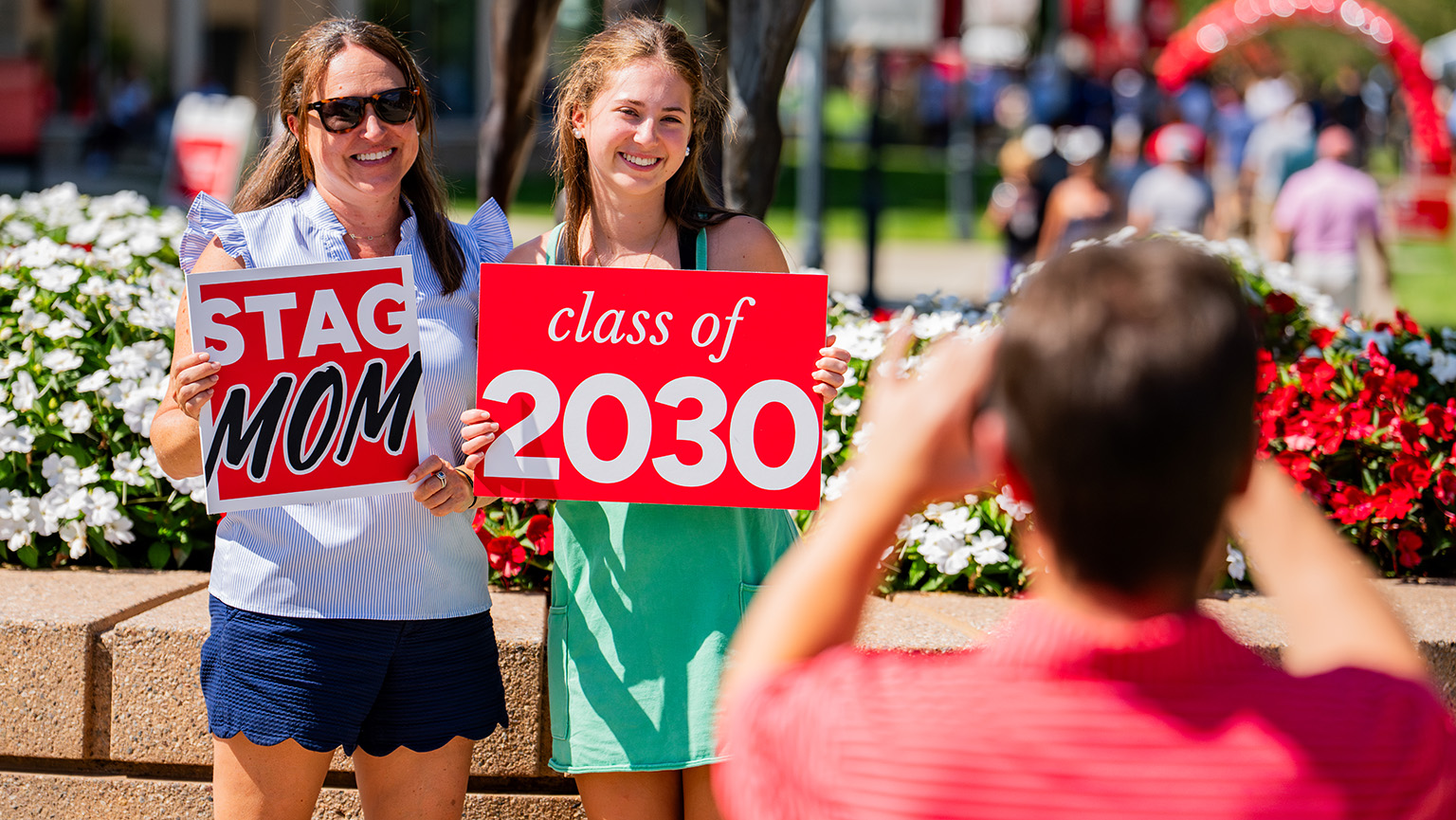 A first-year student holding up a written sign for 'Class of 2030' with her mom (with a written sign for 'Stag Mom') being photographed at move-in.