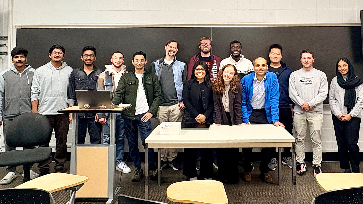 A diverse group of students and instructors poses behind a classroom table and laptop, smiling in a relaxed, welcoming atmosphere.