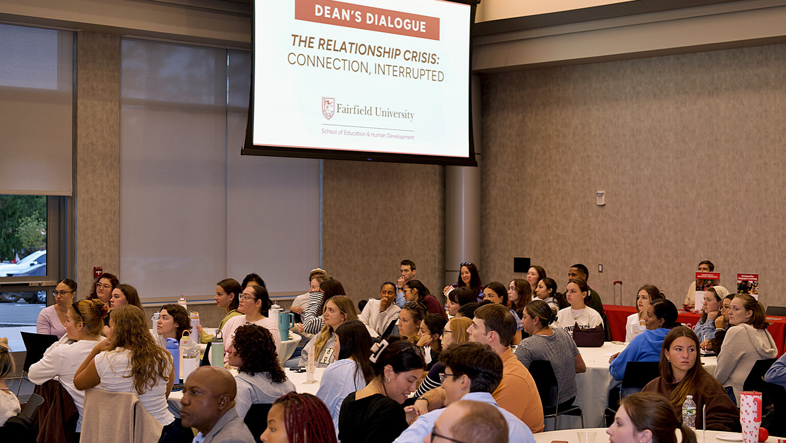 A diverse audience listens in a university lecture hall beneath a screen announcing a Dean’s Dialogue on relationship crises, connection, and interruption.