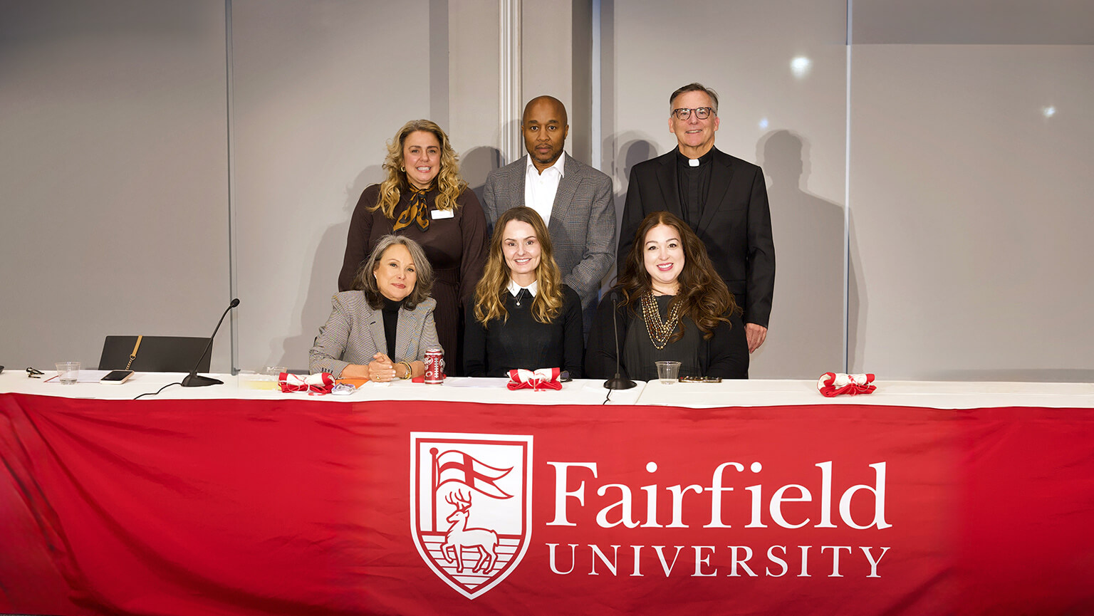 Six people pose behind a table draped with a red Fairfield University banner, smiling at a campus event in a bright, welcoming room.