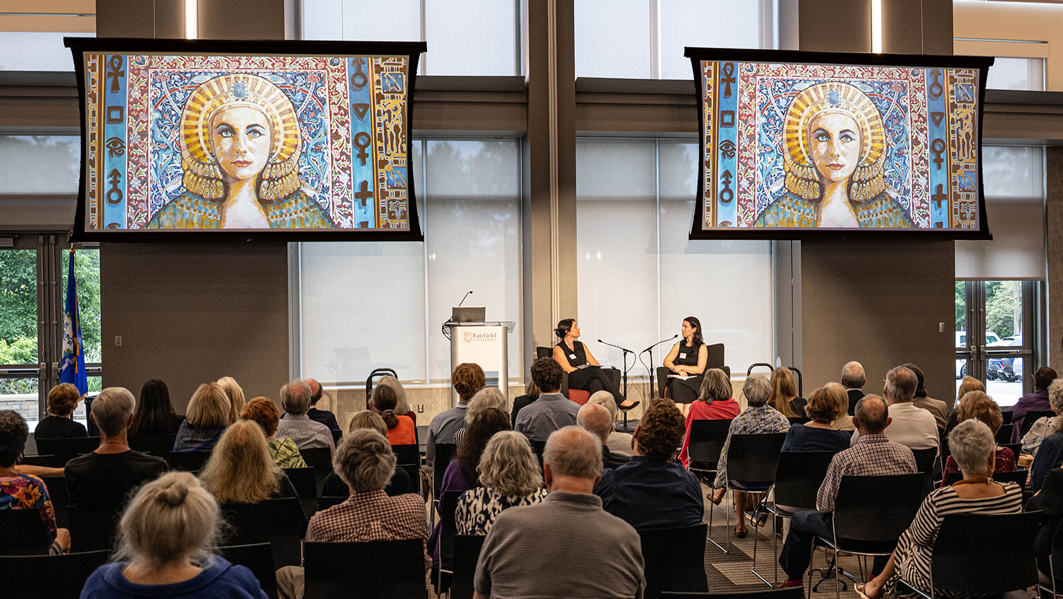 An audience listens to two speakers, Michelle DiMarzo, PhD, and exhibition curator Megan Paqua, at the opening night conversation for art exhibition "Hieroglyphs to Hype: The Modern Afterlife of Ancient Egypt", seated beneath two screens showing colorful portraits of a woman, Audrey Flack’s “Lux Eternal” (2022), creating a thoughtful, reverent atmosphere.
