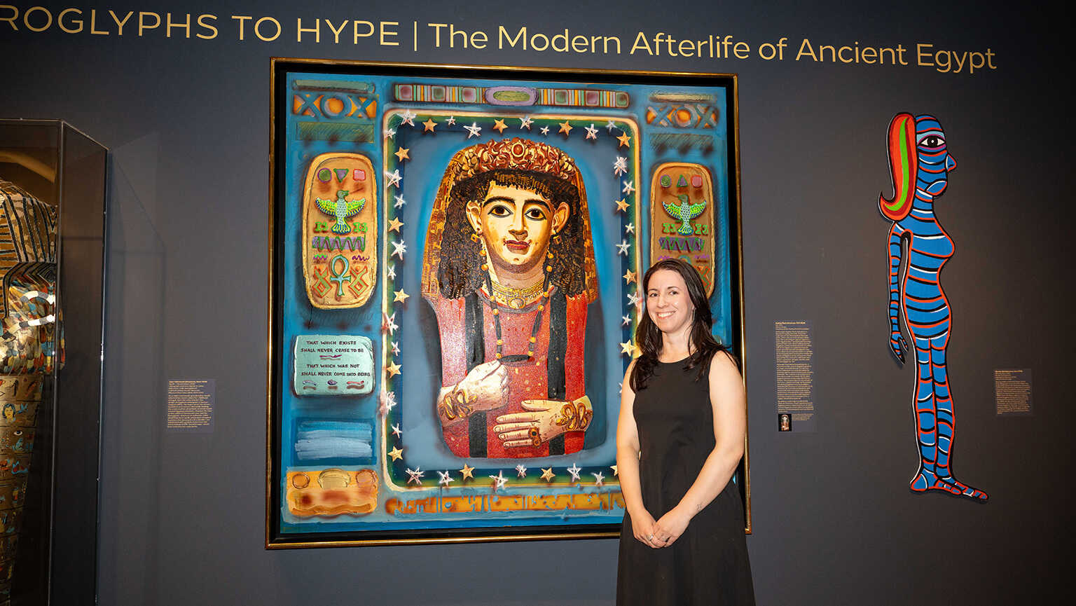 A woman in a black dress, curator Megan Paqua, poses beside a colorful Egyptian-inspired portrait, Audrey Flack’s “Isis” (1983), at “Hieroglyphs to Hype: The Modern Afterlife of Ancient Egypt,” an exhibition at the Fairfield University Art Museum.
