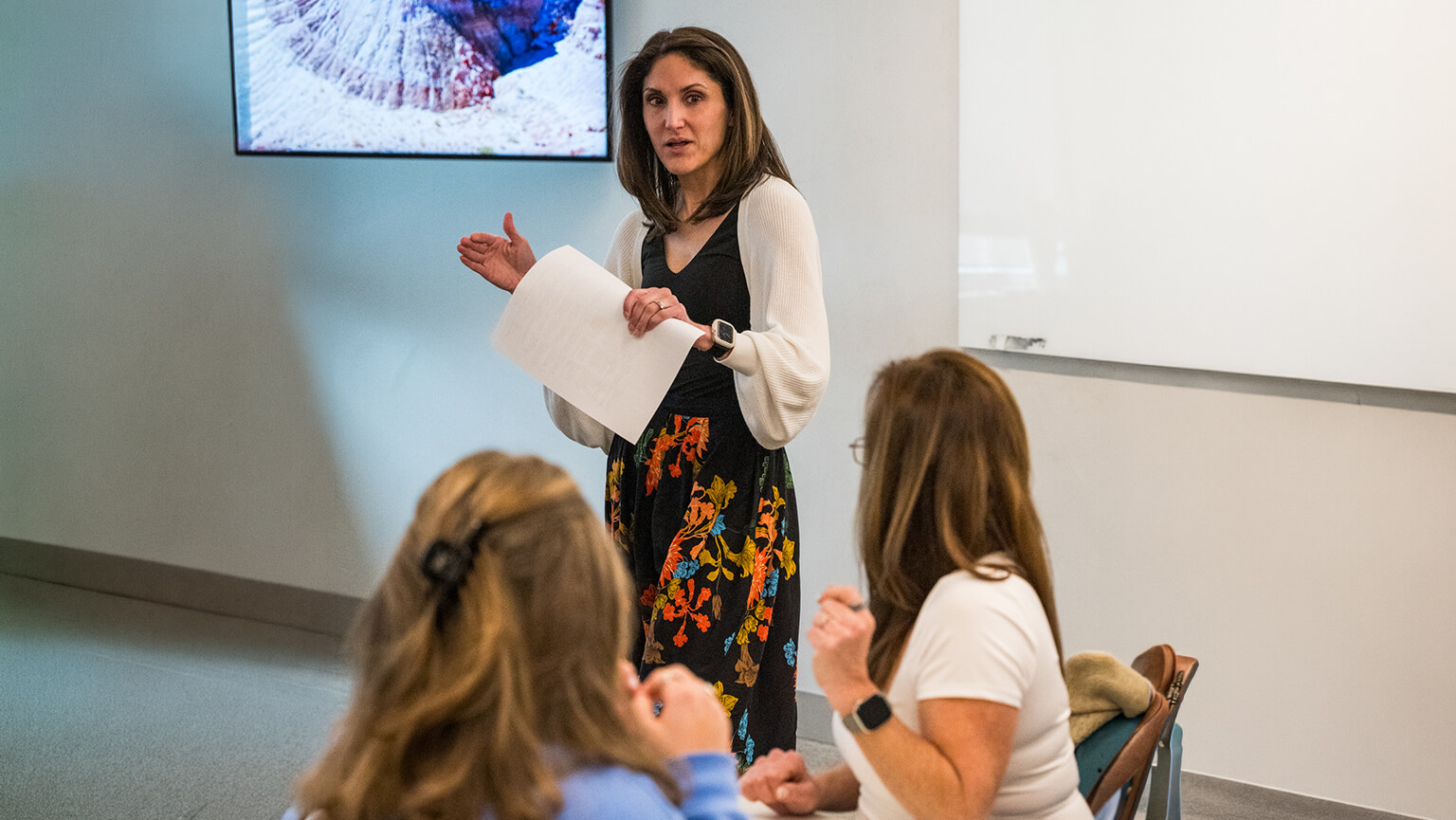 Dr. Kimberly Doughty presents to a small seated audience, holding papers and gesturing beneath a wall-mounted screen; the attentive discussion feels informal and focused.