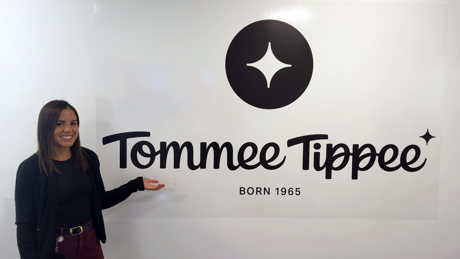 A smiling Mayana Dos Reis MBA’26, MS’26 gestures toward the Tommee Tippee logo on a white wall, with its black star emblem and “Born 1965” text; the scene feels welcoming.