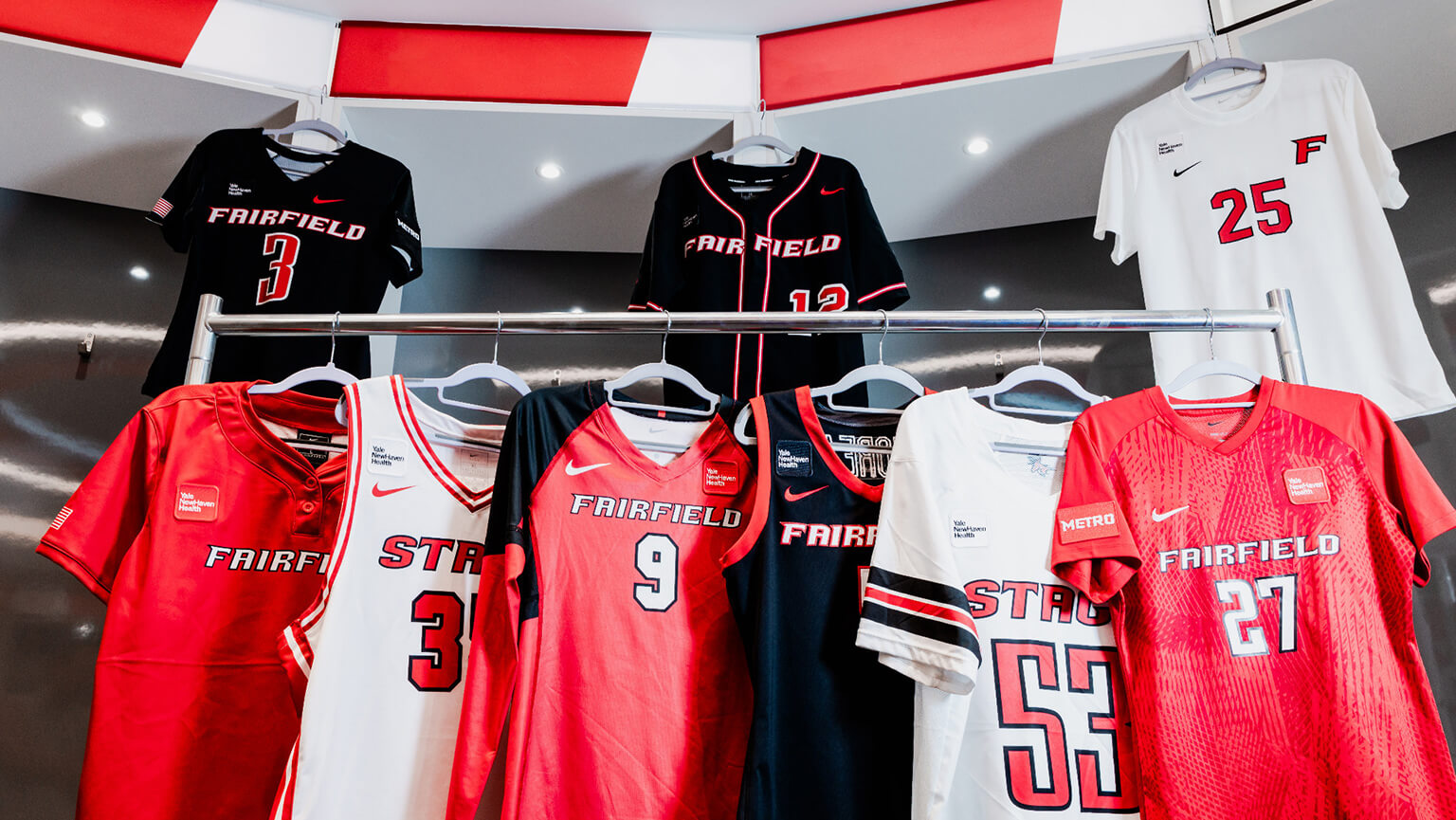A display of Fairfield University sports jerseys on racks, featuring red, black, and white basketball and team shirts with bold numbers and logos.