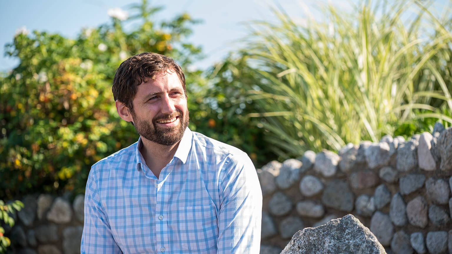 Phil Klay in a blue-and-white checked shirt sits outdoors by a stone wall, with lush greenery behind him in warm sunlight.
