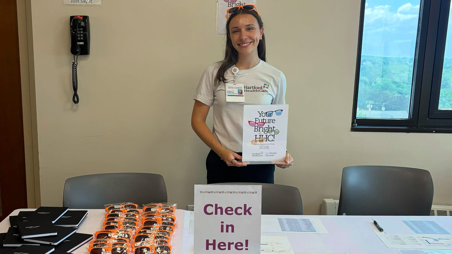 Smiling woman in a white shirt holds a “Your Future Is Bright” certificate behind a “Check in Here!” table lined with brochures and orange items.