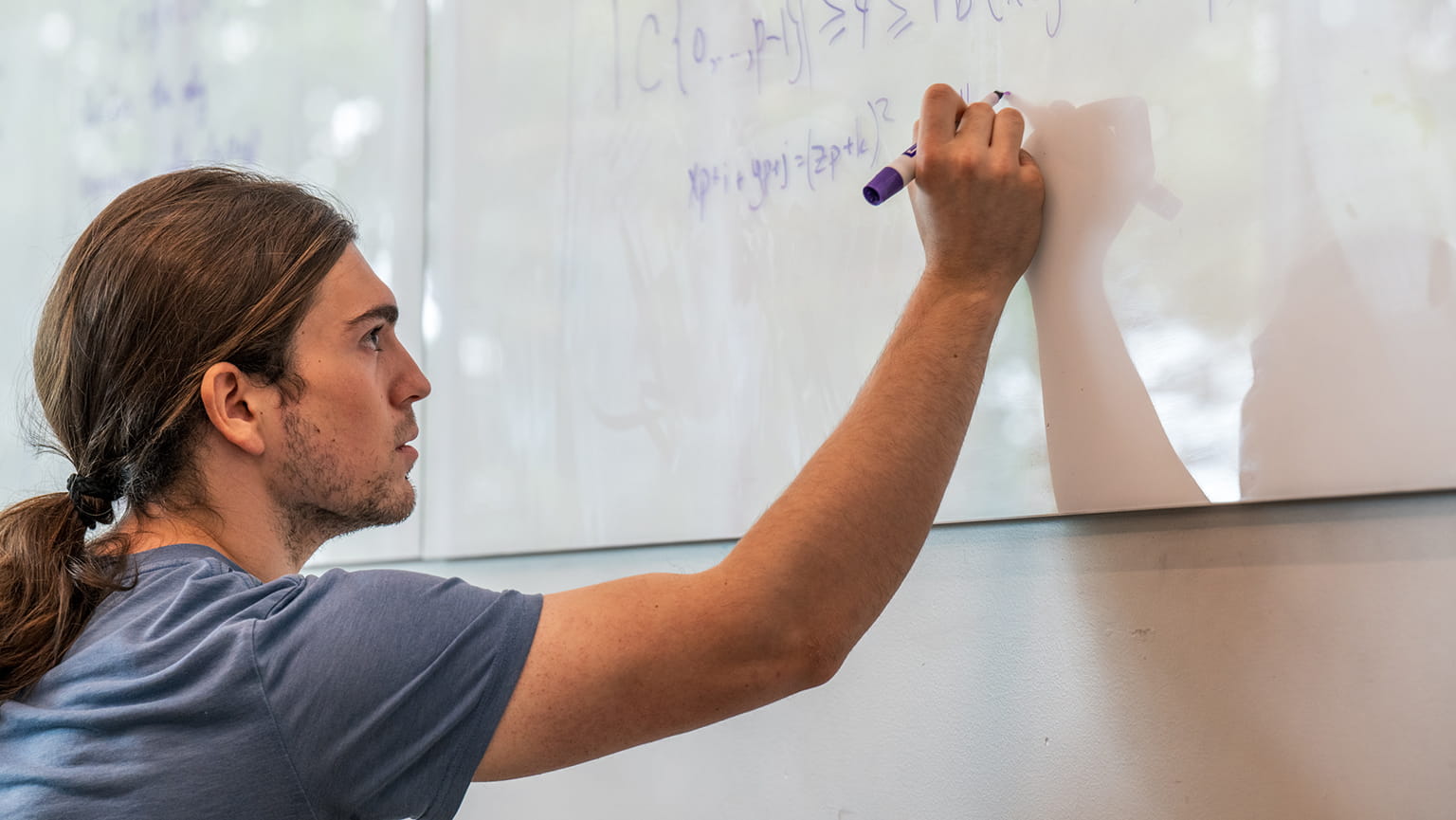 A focused young man with long hair writes equations on a glass whiteboard with a purple marker in a bright classroom, conveying concentration.
