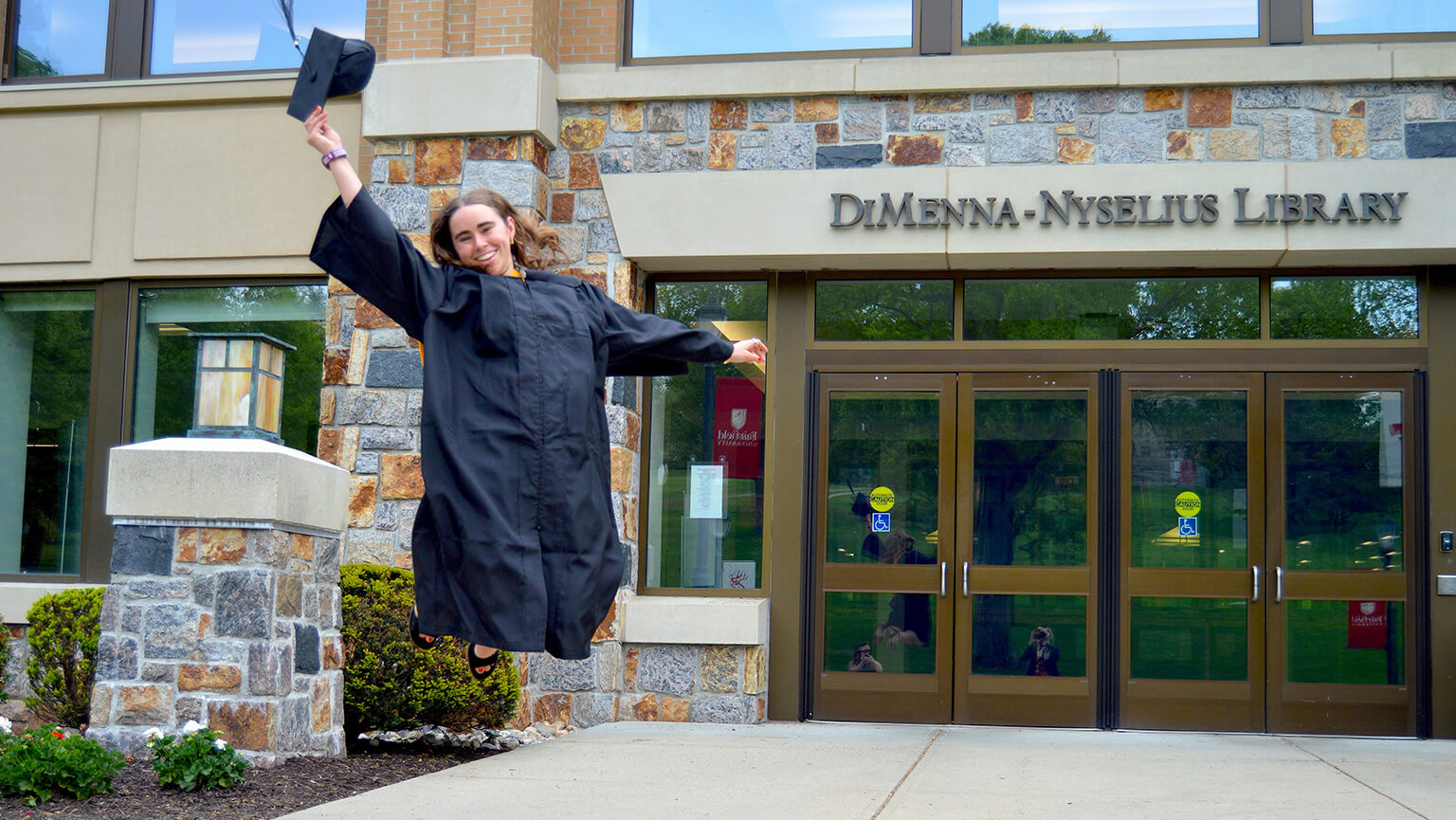 Recent Fairfield Meditz alumna Mia Swenson ’26 in a black gown leaps with one arm raised outside DiMenna-Nyselius Library, celebrating graduation with joyful energy.