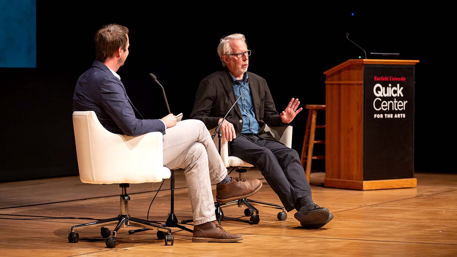 Two men, Andrew Ward and James Welling, converse onstage in white chairs beside a podium labeled “Quick Center for the Arts.” The older speaker, Welling, gestures, creating a thoughtful, formal interview atmosphere.