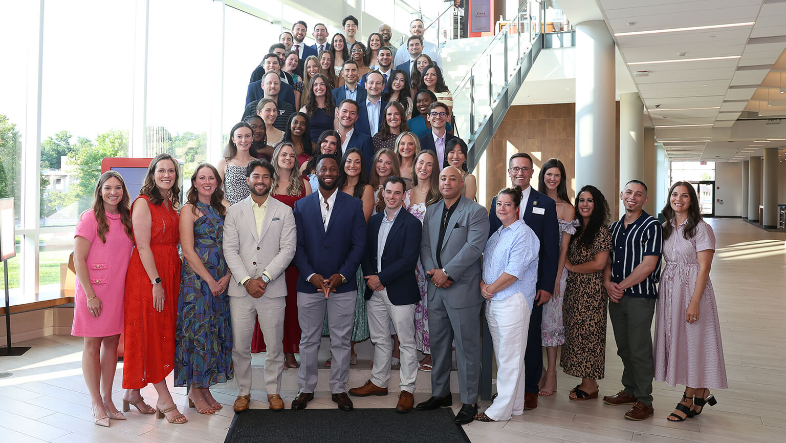 A diverse group of students pose smiling in a bright office atrium beside a staircase, conveying a welcoming, celebratory mood.