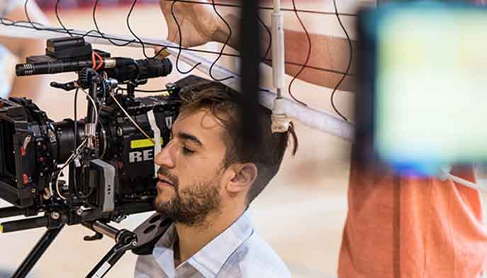 A student operates a camera under a volleyball net.