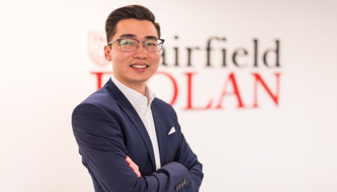 Business student with glasses dressed in a blue suit poses confidently in a branded Fairfield Dolan facility.
