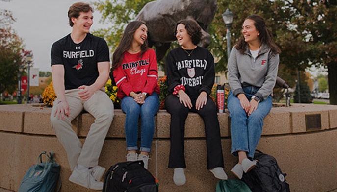 Four students in Fairfield attire, seated before the Stag statue on campus.