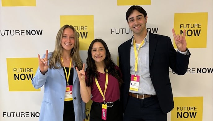 Sophia Haber Brock ’26, Courtney Sabogal ’26, and Justin Sabogal ’25 standing in front of background display showing Future Now logo smiling and waving hands.
