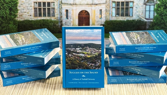 Stacks of the book 'Success on the Sound: A History of Fairfield University' in front of a replica of Bellarmine Hall.