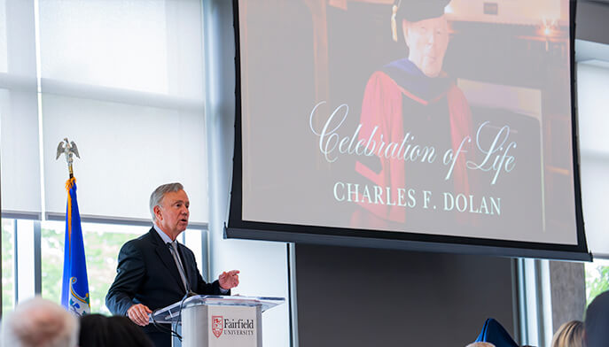 A speaker stands at a podium, addressing an audience with a large screen presentation in the background.