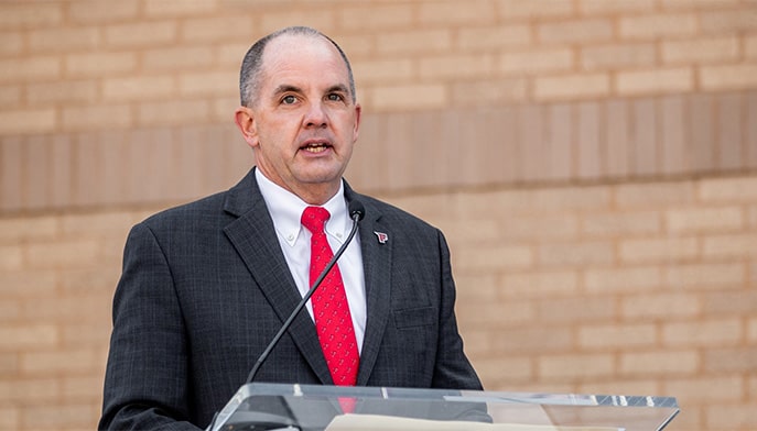 Vice President for Athletics, Paul Schlickmann, giving a speech outdoors from behind a podium.