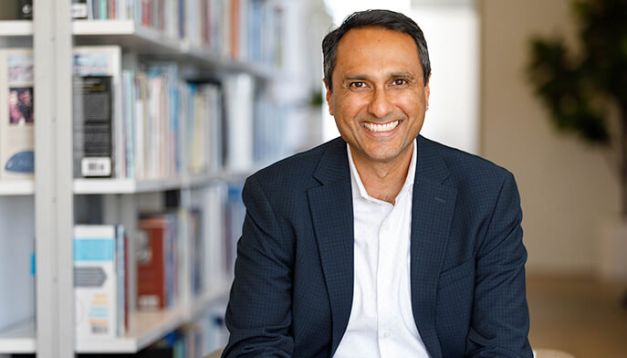 A smiling man in a suit sits in front of bookshelves filled with books, exuding a professional and approachable demeanor.
