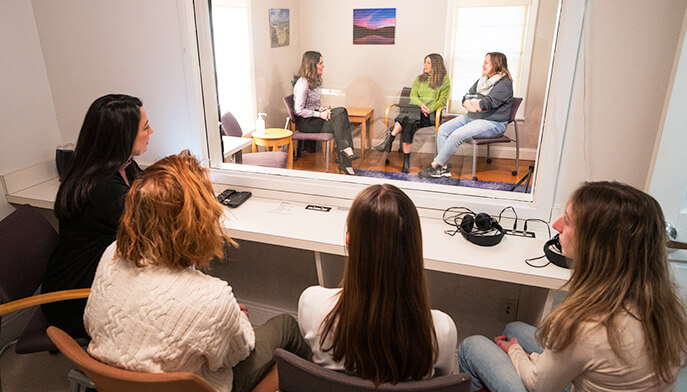 A group of women seated in a room, with a large window reflecting their engaged conversation and expressions.