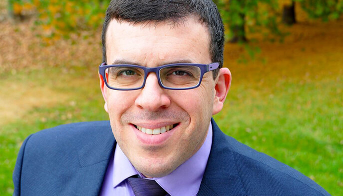 A man in a purple shirt and glasses, standing against a plain background, smiling softly at the viewer.  