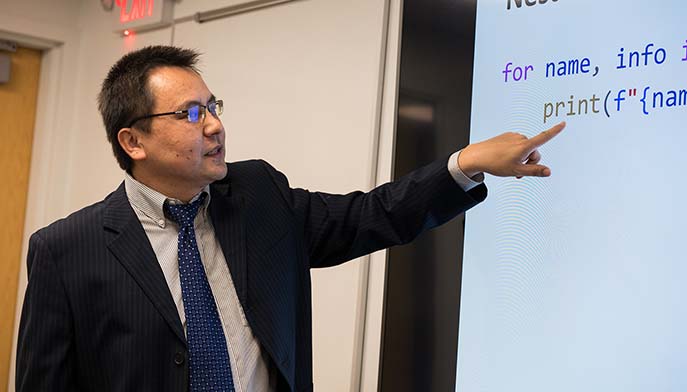 Image of a man in a suit and tie pointing at a screen, likely presenting information to an audience.
