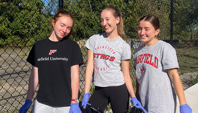 Three girls wearing t-shirts and gloves stand beside a fence, smiling and engaged in an outdoor activity.