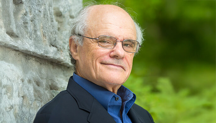 Ken Stein, PhD, with glasses in a blue shirt rests against a stone wall, showcasing a casual pose.