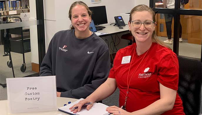 Two women sitting at a table, smiling, with a sign in front of them.