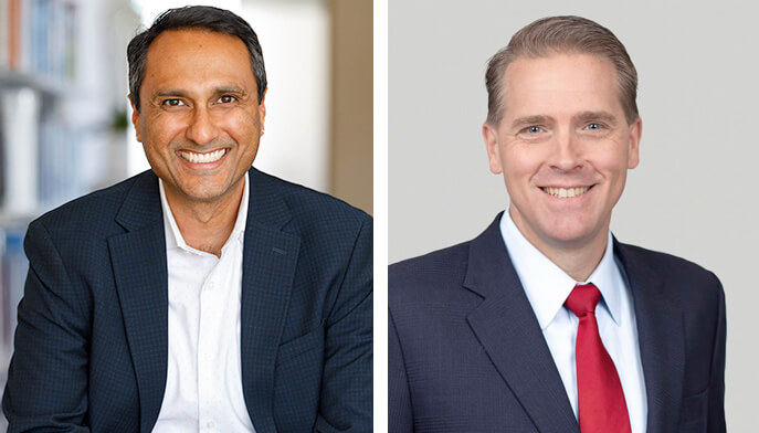 Two men in suits, one smiling broadly and the other wearing a red tie, side by side together in a professional setting.