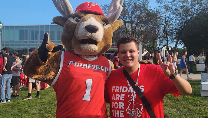 A student poses with Lucas the Stag outside on a bright sunny day on campus.