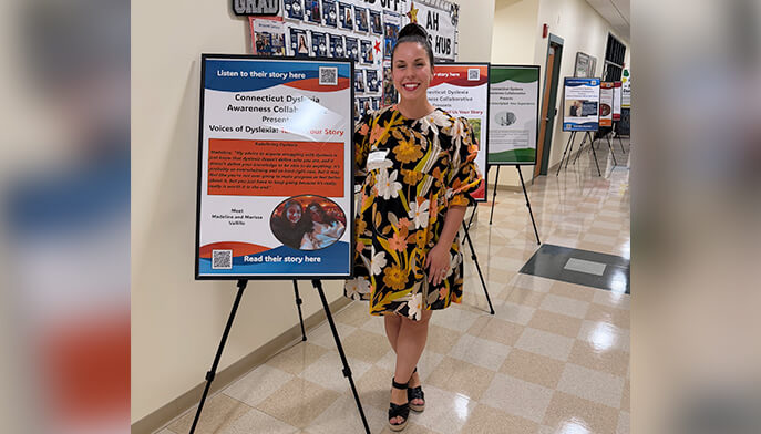 A woman in a dress stands beside a colorful poster, smiling and engaging with the displayed content.