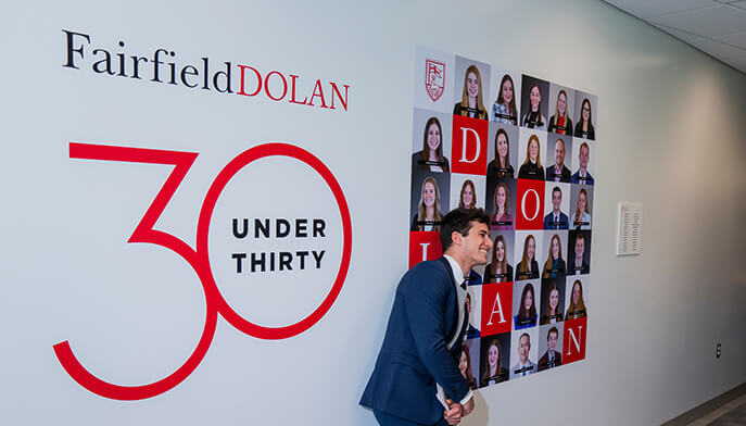 A student dressed in a suit stands before a wall with a prominent sign that reads "30 Under 30," celebrating young achievers.