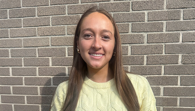 A female student with long brown hair stands in front of a textured brick wall, showcasing her casual pose and expression.  