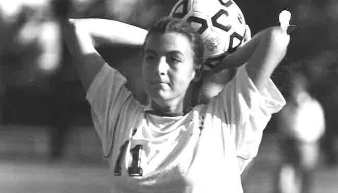 A young woman throwing a soccer ball over her head, smiling confidently in a casual outdoor setting.  