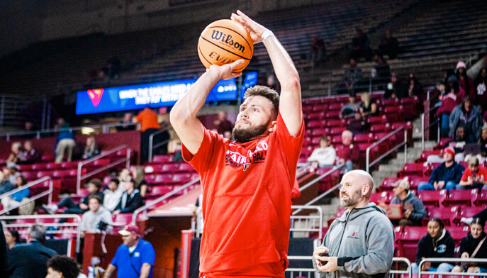 A man student in a red shirt shoots a basketball towards the hoop during a practice.  