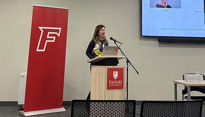 A woman stands at a podium, speaking in front of a large screen displaying information.