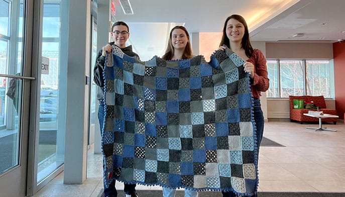 Executive board members of the Stags Stitch comfort shawl program holding up a hand-stitched crocheted quilt.