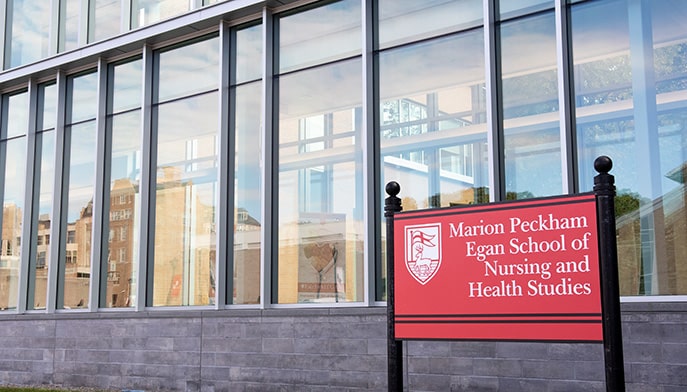 Branded sign for the Marion Peckham Egan School of Nursing and Health Studies in front of its respective glass-window-sided building.
