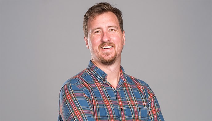 Male professor with blond hair and facial hair smiling for a headshot against a gray backdrop.