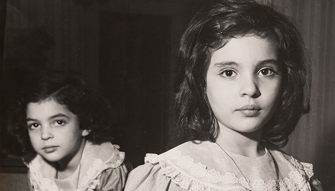 Historic black and white photo of two young girls in frilled dresses wearing heart necklaces.